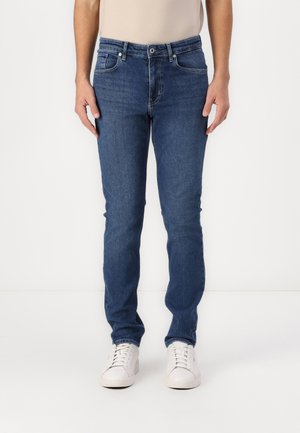 Dark blue denim jeans with a slim fit, five pockets, and a classic waistband, paired with white sneakers on a plain background.