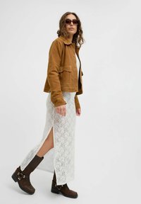 Bruine suede jas met voorzakken, witte maxi jurk van kant met een zijsplit, en zwarte enkelboots met gespen. Bruine zonnebril inbegrepen.