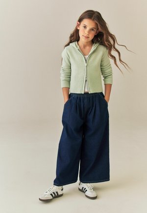 Young girl with long hair wearing a light green zip-up sweater, wide dark blue pants, and white sneakers standing with hands in pockets.