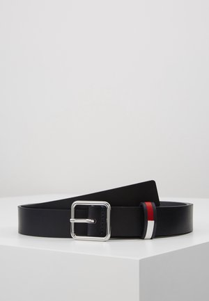 Ceinture en cuir noir avec une boucle rectangulaire argentée et une petite boucle en tissu rouge, blanc et bleu sur une surface blanche.
