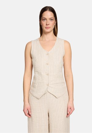 Frau trägt eine beige ärmellose, geknöpfte Weste und passende gestreifte Hose, steht vor einem schlichten hellen Hintergrund.