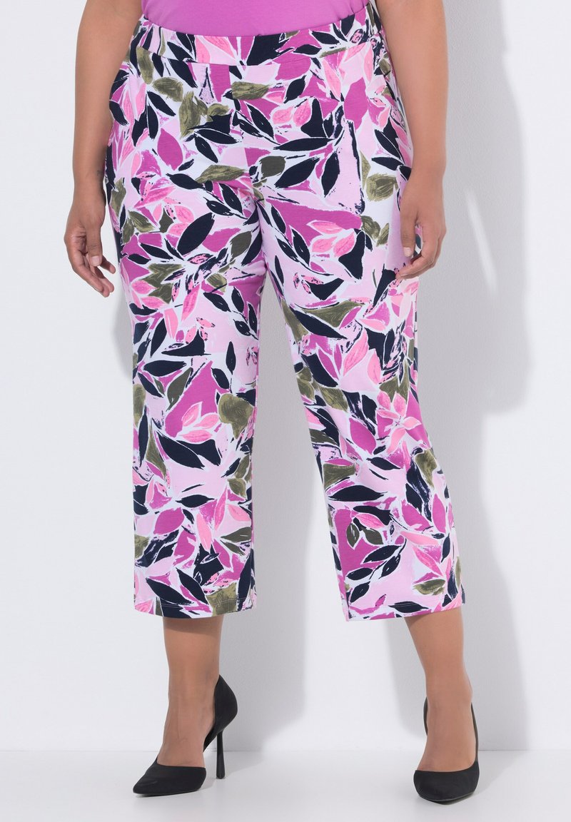 Kobieta w różowo-czarno-zielonych, kwiatowych, krótkich spodniach i czarnych szpilkach na białym tle.