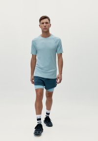 Světle modré sportovní tričko s hladkou texturou, kulatým výstřihem a krátkými rukávy, doplněné o tmavě modré šortky a černé sportovní boty.