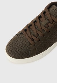 Zapatilla trenzada marrón con un empeine texturizado, cordones marrones oscuros y una suela de goma blanca con un sutil patrón de dibujo.