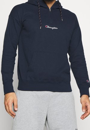 Homme portant un sweat à capuche Champion bleu marine avec poche avant et cordons, associé à un pantalon de survêtement gris clair.