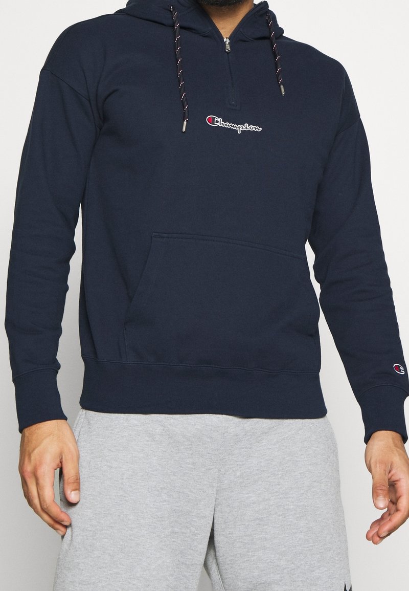Homme portant un sweat à capuche Champion bleu marine avec poche avant et cordons, associé à un pantalon de survêtement gris clair.