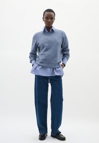 Svijetloplena vuna džemper preko prugaste plave i bijele košulje s ovratnikom, uparen s širokim trapericama i crnim cipelama. Jednostavan, slojevit dizajn.