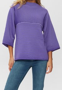 Eine Frau trägt einen lila und lavendelfarben gestreiften Pullover mit drei Viertelärmeln, kombiniert mit blauen Jeans vor einem schlichten Hintergrund.