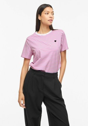 Femme portant un t-shirt rayé rose et blanc avec un petit cœur noir sur la poitrine et un pantalon noir taille haute, debout avec une main dans la poche.