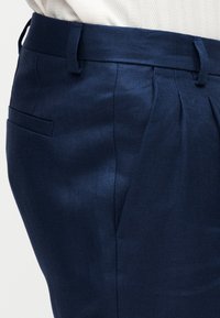 Σκούρο μπλε σμόκιν παντελόνια με θηλιές για ζώνη και πλαϊνή τσέπη, φορεμένα με λευκό υφασμάτινο τοπ.