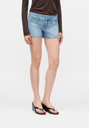 Žena v svetlomodrých rifľových kraťasoch, hnedom tričku s dlhým rukávom a čiernych klinových žabkách, stojí pred obyčajným pozadím.