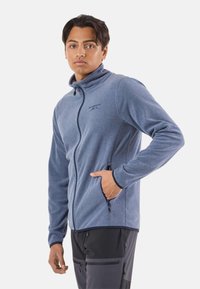 Flis jakna svijetloplave boje s potpunim patentnim zatvaračem, visokim ovratnikom i bočnim džepovima. Ima kontrastne crne manžete i patentne zatvarače.