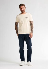 Kermaisen värinen t-paita, jossa on 'PTRL'-teksti, yhdistetty tummansinisiin farkkuihin. Mallilla on valkoiset tennarit. Kangas vaikuttaa pehmeältä ja rentoon tyyliin sopivalta.