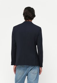 Man med mörkt hår klädd i en figurnära svart kavaj och ljusblå jeans, stående med ryggen mot kameran mot en vit bakgrund.