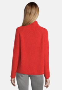 Maglione rosso lavorato a maglia con collo alto, maniche raglan e finitura strutturata. Presenta una vestibilità ampia e dettagli a coste attorno al colletto.