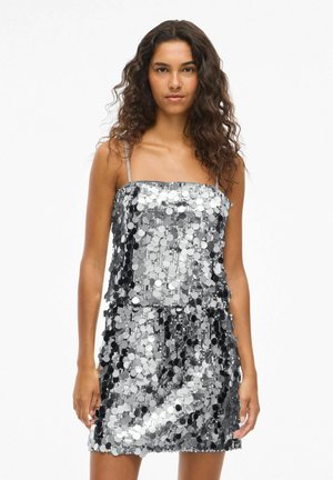 Srebrna mini obleka s paljetami in tankimi naramnicami, ki ima valovito teksturo in različne odtenke srebrne in črne. Rob je neenakomeren.