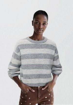 Maglione a righe grigio e crema con scollo a giro e maniche arrotolate, abbinato a pantaloni marroni con borchie argentate lungo le tasche.