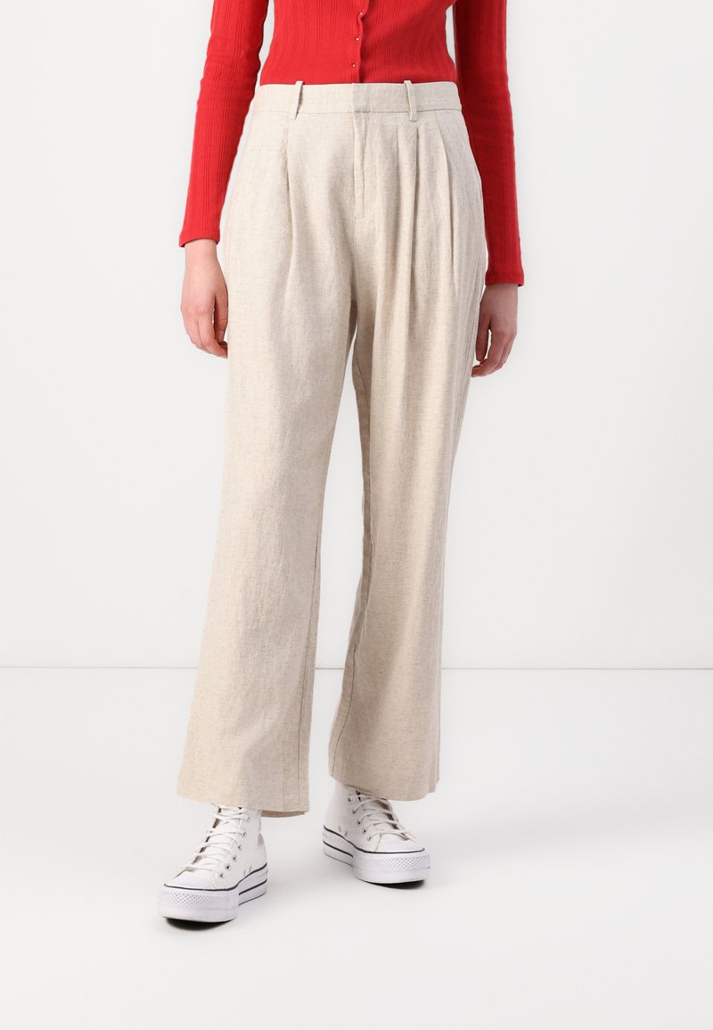 Pantalons plissés beiges à coupe ample, fabriqués en tissu texturé, associés à des baskets blanches et un haut rouge ajusté.