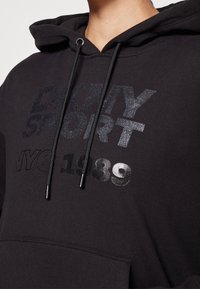 Sweat à capuche noir avec texte brillant "DKNY Sport NYC 1989" et cordons noirs, porté par une personne cadrée au niveau du cou et de la poitrine.