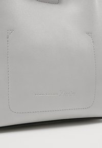 Surface en cuir gris clair avec un patch rectangulaire cousu et un petit texte en relief indiquant "Tom Tailor Denim" près du centre en bas.