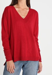 Maglione rosso a scollo a V con maniche lunghe e spacchi laterali. Presenta dettagli con bottoni sui polsini delle maniche. Realizzato in materiale morbido e leggero.