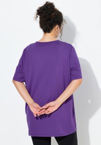 Fioletowy krótki t-shirt z gładkiej tkaniny, o luźnym kroju i zaokrąglonym dekolcie, z rękami umieszczonymi za plecami.