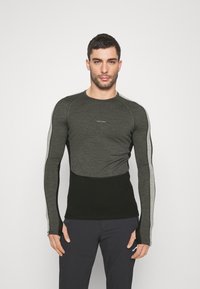 Mężczyzna w dopasowanej, długiej koszulce sportowej w dwóch odcieniach szarości i czerni oraz ciemnych spodniach, stojący na tle jednolitego białego tła.