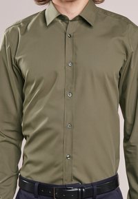 Camisa de botones color verde oliva con textura suave, que presenta un cuello clásico y botones negros, combinada con un cinturón oscuro y pantalones azul marino.