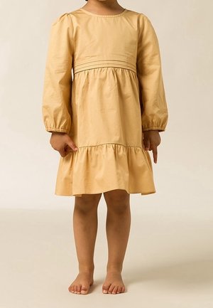 Kind draagt een lange mouw beige jurk met een geplooide taille en zoom, staat blootsvoets tegen een neutrale achtergrond.