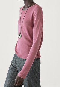 Pull en maille côtelée à manches longues de couleur rose pâle. Associé à un pantalon rayé sombre. Un collier pendentif avec un accent circulaire complète le look.