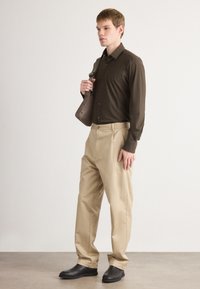 Camisa marrón de manga larga con botones, pantalones beige con pliegues y zapatos negros sin cordones. El modelo lleva un pequeño bolso de hombro.