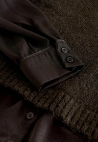 Dark, textured knit sweater overlaps a smooth, dark fabric shirt with buttoned cuff. The colors are muted, primarily shades of brown and green.
