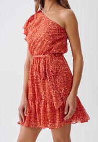 Femme portant une robe sans manches, une épaule, à motifs rouges et oranges, avec une ceinture nouée à la taille et un ourlet à volants.