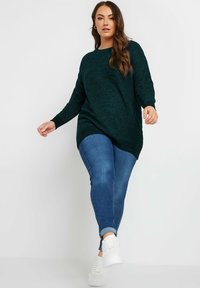 Donkergroene, textuurgebreide trui met een losse pasvorm, ronde halslijn en lange mouwen, gecombineerd met blauwe skinny jeans en witte sneakers.