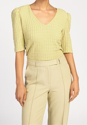 Kobieta w jasnżółtej, fakturowanej bluzce z dekoltem w serek, z rękawami do łokci i wysokim stanem beżowych, dopasowanych spodni, stojąca na jednolitym tle.