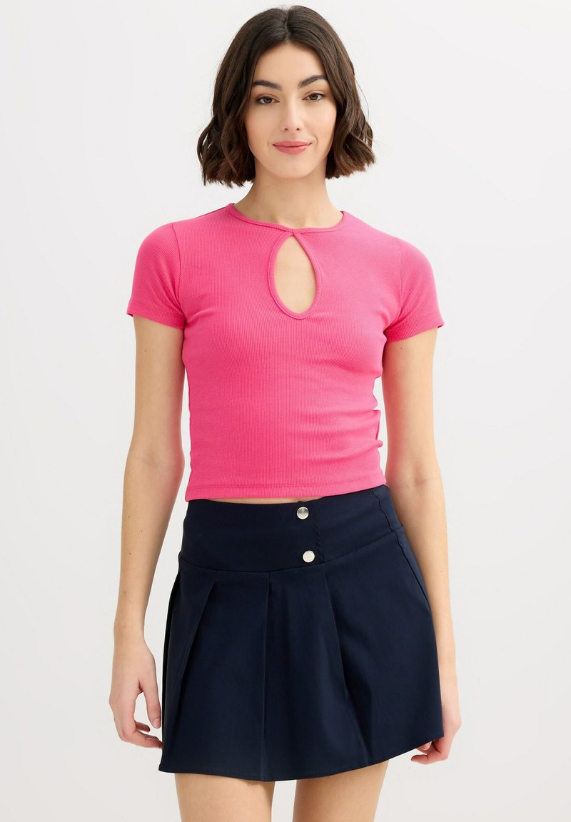 Mujer joven con cabello corto y oscuro, vistiendo una blusa de manga corta de color rosa brillante con una abertura frontal y una falda plisada azul marino de talle alto.