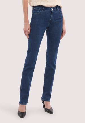 Dunkelblaue Slim-Fit-Jeans kombiniert mit schwarzen spitzen High Heels und einem hellen, gemusterten Oberteil, das in der Hose steckt.