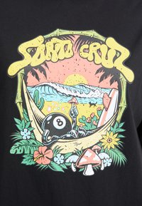 T-shirt nera con una vivace grafica. Immagine centrale di un personaggio a forma di palla 8 in un'amaca con un tramonto, onde e piante tropicali.