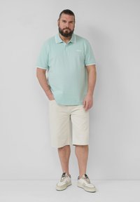 Hellgrünes Polohemd mit weißen Akzenten, kombiniert mit beigen Shorts und cremefarbenen Sneakern. Einfaches Design, Baumwollmaterial, lässige Passform.