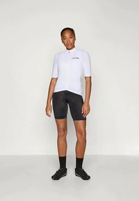 Λευκή ποδηλατική μπλούζα με κοντά μανίκια και φερμουάρ μπροστά, συνοδευόμενη από μαύρο λογότυπο, συνδυασμένη με μαύρες σορτς με επένδυση. Το μοντέλο φοράει μαύρα ποδηλατικά παπούτσια και κάλτσες.