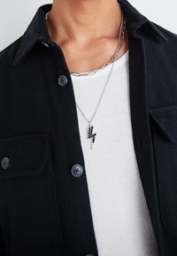 Svart skjorta med knappar över en vit ribbad T-shirt, prydd med lager av silverkedjor. En silverblixtpendel med svarta detaljer.