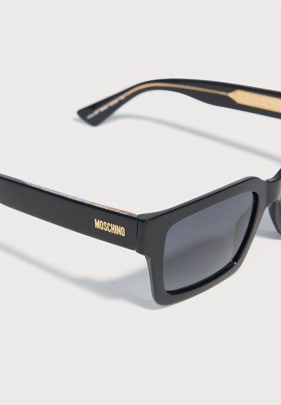 Μαύρα ορθογώνια γυαλιά ηλίου Moschino με σκούρους φακούς και χρυσή σήμανση στο πλάι, παρουσιάζονται σε λευκό φόντο.