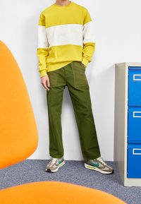 Osoba v žluto-bílé mikině Nike, olivově zelených kalhotách a šedých teniskách s tyrkysovým logem stojí vedle modrého šanonu.