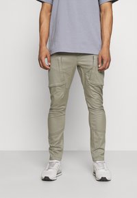 Ljus olivgröna cargo-byxor med flera fickor, smal passform och texturerad tyg. Bärs med en ljusgrå t-shirt och vita sneakers.