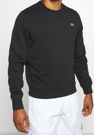 Homme portant un sweat-shirt noir à manches longues avec un petit logo vert sur la poitrine et un short de sport blanc, sur fond uni.