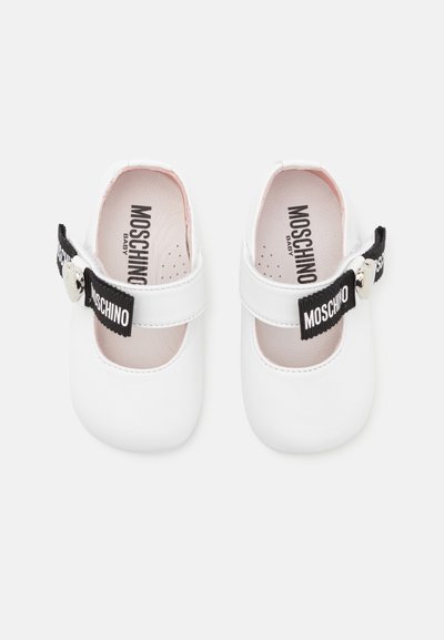 Bílé dětské boty vyrobené z hladkého materiálu. Obsahují černý pásek s textem "MOSCHINO" a stříbrnými doplňky. Zaoblený design špičky.