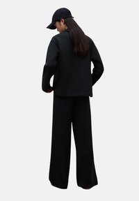 Persona con cabello largo, usando una gorra de béisbol negra, una camiseta de manga larga negra y pantalones anchos negros, de pie contra un fondo liso.