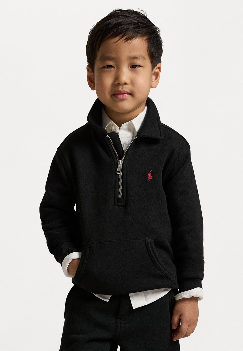 Schwarze Zip-up-Sweatjacke aus strukturiertem Stoff mit einem roten Logo auf der Brust, kombiniert mit einem weißen Hemd mit Kragen darunter.