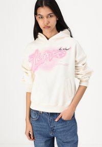 Felpa crema con design grafico rosa che presenta la parola "lights". Materiale morbido, tasca a marsupio e polsini a costine. Indossata con jeans blu.