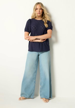 Blonde Frau steht mit verschränkten Armen, trägt ein marineblaues kurzärmeliges Oberteil mit Lochstickerei, weite hellblaue Jeans und weiße Sandalen.